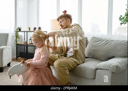 Vater mit verrückter Frisur und verrücktem Gesicht Grimace, der seiner Tochter Haare macht, glückliche Eltern Stockfoto