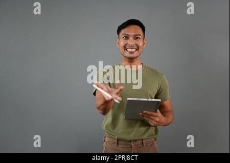 Ein lächelnder asiatischer Millennials in lässiger Kleidung mit seinem digitalen Tablet steht isoliert auf grauem Studiohintergrund mit einer Geste zur Präsentation. Stockfoto