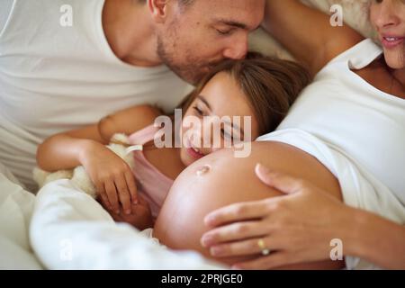 Und bald werden es vier sein. Ein glückliches kleines Mädchen, das mit ihrem Vater und ihrer Schwangeren im Bett liegt. Stockfoto