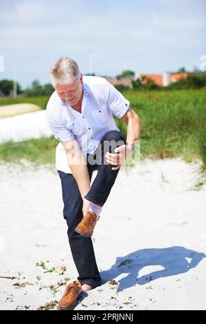 Er will den Strand unter seinen Füßen spüren. Ein älterer Mann, der seine Schuhe am Strand auszieht Stockfoto