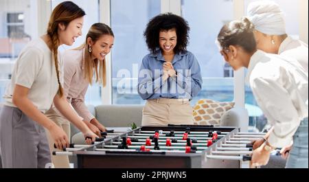 Teambuilding-, Break- und Geschäftsfrauen, die in der Bürolounge spielen, Spaß haben und sich zusammentun. Trendige, coole und vielfältige kreative Mitarbeiter mit lustigen Mini-Tischfußballspielen Stockfoto