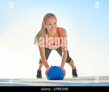 Ich finde so viel Spaß in Fitness. Ganzkörperportrait einer jungen Frau, die Liegestütze mit einem Medizinball macht. Stockfoto