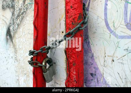 Ein altes Vorhängeschloss auf einer rostigen Kette, die um rote Rohre gewickelt ist. Stockfoto