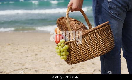 Ein Mann hält einen Korbkorb mit Essen und Wein für ein Picknick am Strand. Sommerliches Entspannungs- und Erholungskonzept. Stockfoto