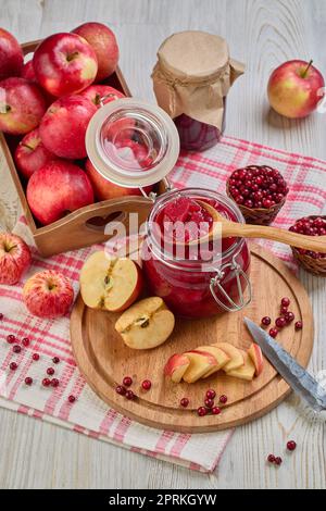 Stillleben mit frischen Preiselbeeren, roten Äpfeln mit Messer auf Holzbrett und Glasbecher mit hausgemachter Marmelade auf hellem Hintergrund. Herbsternte, Stockfoto