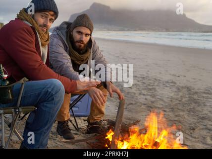 Es ist kälter, als es aussieht. Zwei junge Männer, die am Strand am Feuer sitzen Stockfoto