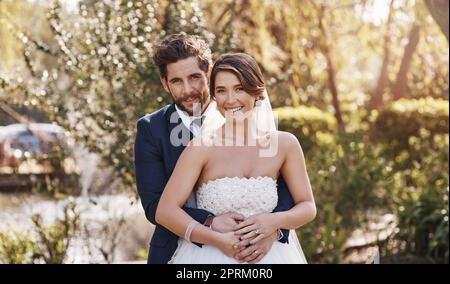 Wir haben uns entschieden, uns zu verpflichten. Gekürztes Porträt eines liebevollen jungen Bräutigams, der seine Braut von hinten umarmt, während er auf seiner Hochzeit draußen steht Stockfoto