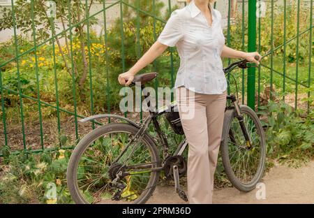 Im mittleren Alter attraktive schlanke Frau in leichte Hose und Hemd steht in der Nähe der Fahrrad im Park an einem sonnigen Sommertag, Radfahren Konzept für erwachsene Frauen. Stockfoto