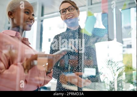 Der Plan sieht idiotensicher aus. Zwei Geschäftsfrauen, die in einem Büro mit Notizen an einer Glaswand Brainstorming betreiben Stockfoto