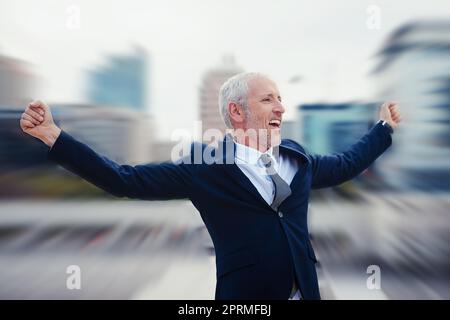 Es ist ein toller Tag, um geschäftlich zu sein. Verbesserte Aufnahme eines glücklichen Geschäftsmannes, der seine Arme hebt, während er in der Stadt steht. Stockfoto