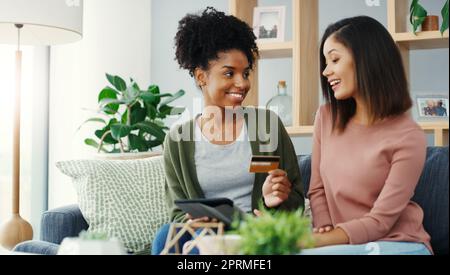 Sollten wir online einkaufen? Zwei attraktive junge Frauen, die zusammen ein Tablet zum Online-Shopping nutzen, während sie auf dem Sofa sitzen. Stockfoto