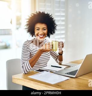 Sie hat eine strahlende Zukunft vor sich. Porträt einer fröhlichen jungen Frau, die an einem Laptop arbeitet und Kaffee trinkt, während sie zu Hause auf die Kamera schaut. Stockfoto