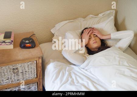 Warum kann ich heute nicht einfach im Bett bleiben? Eine junge Frau wacht morgens zu ihrem Wecker auf. Stockfoto