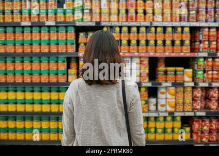 Vergleich aller Marken und Preise. Rückansicht einer Frau, die in einem Lebensmittelgeschäft nach Gegenständen browsen soll. Stockfoto