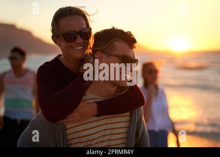 Verliebt unter der Dämmerung. Ein liebevoller junger Mann, der seine Freundin am Strand mit dem Huckepack betreibt Stockfoto