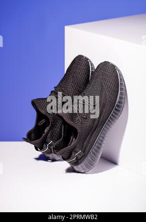 Schwarze Sneaker auf blauem Hintergrund mit weißem Podium in Form eines geometrischen Würfels. Stockfoto