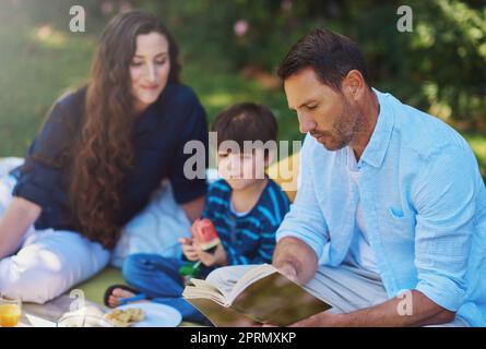 Jede Familie hat eine Geschichte. Eltern lesen ihrem Sohn während eines Picknicks im Park ein Buch vor. Stockfoto