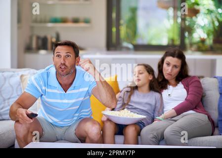 Ein Mann, der Sport im Fernsehen sieht, während seine Familie im Hintergrund gelangweilt sitzt. Stockfoto