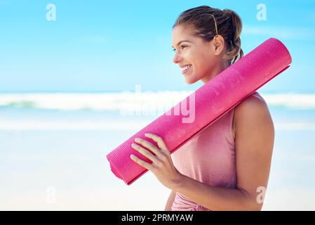 Der perfekte Tag für Yoga im Freien. Eine junge Frau, die eine Yogamatte hält, während sie am Strand steht. Stockfoto