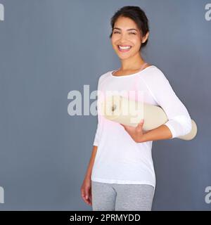 Starten Sie heute. Eine sportliche junge Frau, die ihre Yogamatte vor einem grauen Hintergrund hält. Stockfoto