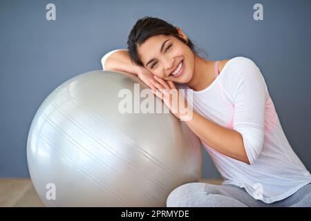 Nehmen Sie sich Zeit, um zu tun, was Ihre Seele glücklich macht. Eine sportliche junge Frau, die sich vor grauem Hintergrund auf einen Pilates-Ball stützt. Stockfoto