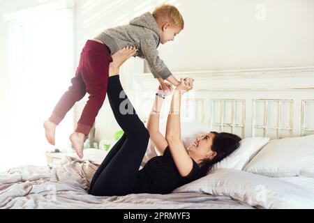 Hoch mit dir. Ein fröhlicher kleiner Junge, der tagsüber von den Beinen seiner Mutter im Schlafzimmer hochgehoben wird Stockfoto