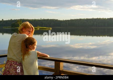 Mutter und Tochter, die sich im Abendlicht auf ein Geländer neben einem See lehnen Stockfoto