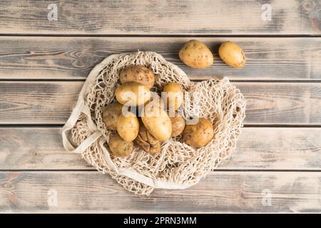 Haufen ungeschälter gelber neuer Kartoffeln auf verblichenem Holztisch, Draufsicht. Frisches Bauerngemüse in Öko-Stoffbeutel. Bio-Lebensmittel oder gesunde Ernährung Konz Stockfoto