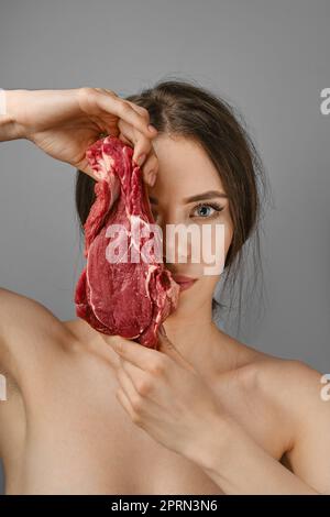 Die junge Frau hält rohes Rindfleisch und bedeckt die Hälfte ihres Gesichts Stockfoto