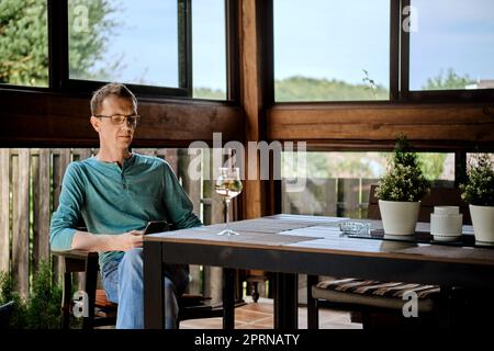 Ein Mann mittleren Alters sitzt auf der Veranda des Landhauses mit einem Glas Wein und benutzt ein Smartphone Stockfoto