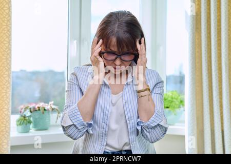 Reife Frau hat Kopfschmerzen, hält den Kopf in der Hand, steht neben dem Fenster Stockfoto