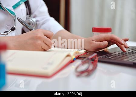 Eine Ärztin, die im Büro arbeitet, sich Notizen im Tagebuch macht und auf dem Laptop tippt Stockfoto