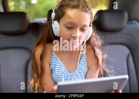 Ein junges Mädchen, das in einem Auto auf dem Rücksitz sitzt und Kopfhörer trägt und ein digitales Tablet benutzt. Stockfoto