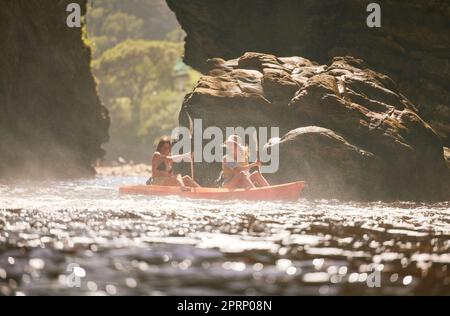 Reisen, Kajak und weibliche Freunde auf einem Urlaubsabenteuer auf einem Fluss oder Meer mit Blick auf die Berge in der Natur. Frauen, die Spaß auf Reisen haben und Kanufahren auf dem Urlaub genießen Stockfoto