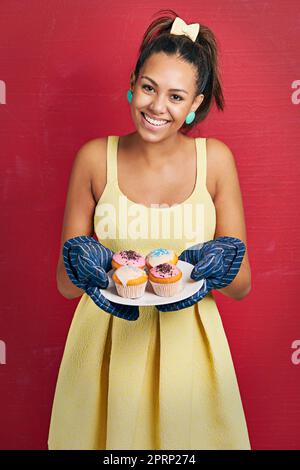 Frisch aus dem Ofen. Studioaufnahme einer jungen Frau, die einen Teller Cupcakes vor einem roten Hintergrund hält. Stockfoto