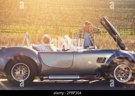 Dieser Roadtrip hat einen sehr schlechten Start. Ein älterer Mann schaut unter die Motorhaube seines Autos, während er mit seiner Frau unterwegs ist. Stockfoto