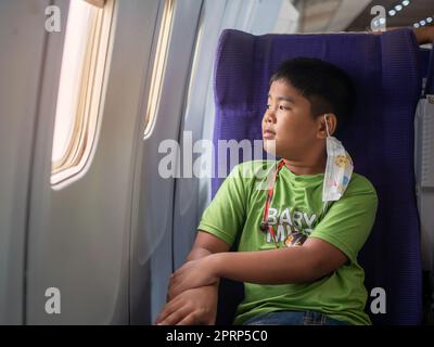 Ein asiatischer Junge sitzt und lächelt und schaut aus dem Fenster eines Flugzeugs. Stockfoto