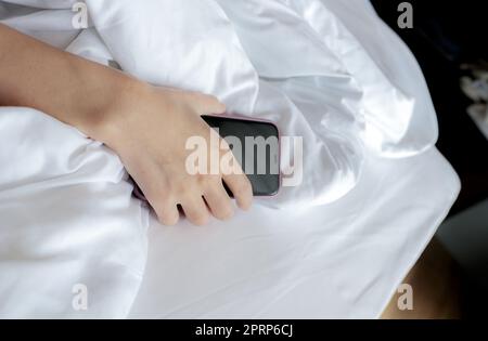 Frau schläft im Bett und Hand hält Handy. Frau, die im Schlafzimmer ein Smartphone benutzt. Eine Frau, die Smartphone benutzt. SMS auf dem Handy mit Müdigkeit bis zum Einschlafen. Sie schläft mit dem Smartphone. Stockfoto