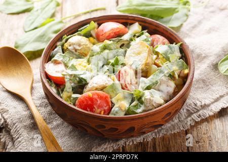 Köstlicher Vitaminsalat mit gekochten Eiern, Kartoffeln, Tomaten, Zwiebeln und Sorrel, Spinat mit Joghurt aus der Nähe in einer Schüssel auf einem Holztisch. Hori Stockfoto