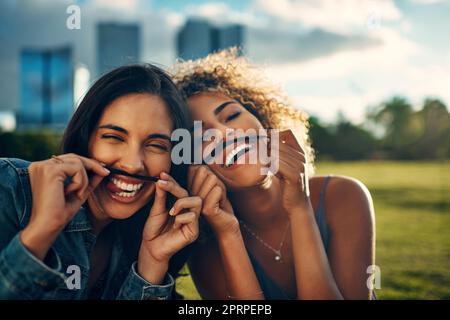 Wir lieben es, zusammen albern zu sein. Verkürztes Porträt von zwei attraktiven jungen Freundinnen, die mit ihren Haaren einen Schnurrbart in einem Park machen Stockfoto