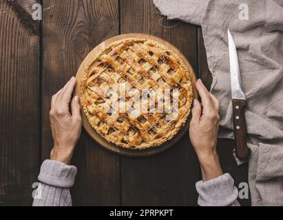 Zwei weibliche Hände halten einen runden gebackenen Kuchen mit Apfelfüllung auf einem Holzbrett, braunem Tisch. Stockfoto