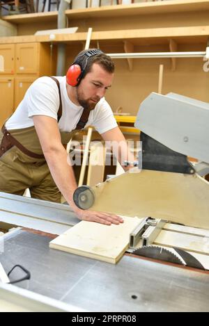 Junger Zimmermann in Arbeitskleidung arbeitet in der Schreinerei an einer Schleifmaschine - Arbeitskleidung mit Gehörschutz Stockfoto