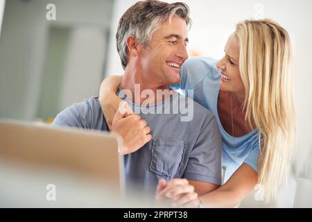 Sie bedeutet mir die Welt. Eine attraktive reife Frau umarmt ihren hübschen reifen Mann, während er im Haus einen Laptop benutzt. Stockfoto