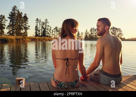 Genießen Sie die letzten Strahlen eines Sommertages. Ein liebevolles junges Paar in Badeanzügen, das bei Sonnenuntergang auf einem Dock sitzt. Stockfoto