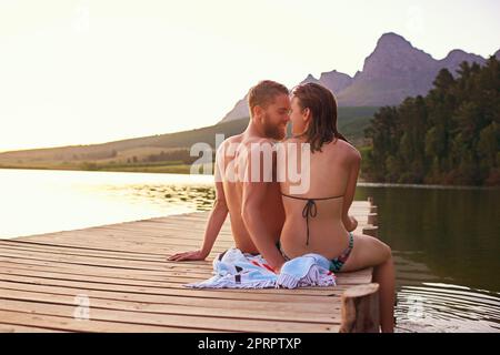 Den Tag im Leerlauf verbringen. Rückansicht eines liebevollen jungen Paares in Badeanzügen, das bei Sonnenuntergang auf einem Dock sitzt. Stockfoto