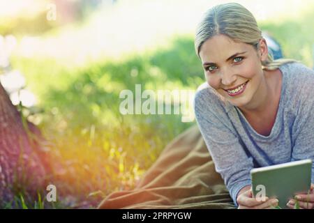 WLAN überall. Porträt einer schönen jungen Frau, die auf dem Gras liegt und ein digitales Tablet benutzt. Stockfoto