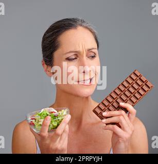 Es ist eine wirklich schwierige Entscheidung. Studioaufnahme einer attraktiven reifen Frau, die sich zwischen Salat und Schokolade entscheidet. Stockfoto