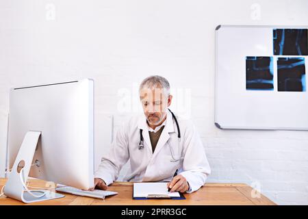 Aufzeichnen von Patientendaten. Ein reifer Arzt, der an einem Desktop-Computer in seinem Büro arbeitet. Stockfoto