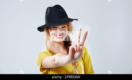 Den Frieden teilen. Beschnittenes Studioporträt einer schönen jungen Frau mit schwarzem Hut und Friedenszeichen. Stockfoto