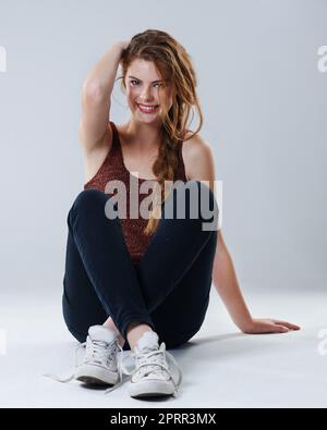 Shes hat Stil und die richtige Einstellung. Porträt einer schönen jungen Frau in Casualwear, die mit gekreuzten Beinen auf dem Studioboden sitzt. Stockfoto